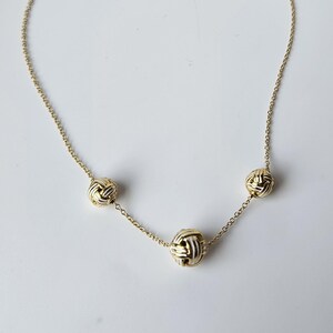 May include: A gold-toned necklace featuring three decorative knot-shaped pendants. The necklace has a delicate chain and the pendants have a woven design. The jewelry is displayed on a white background.