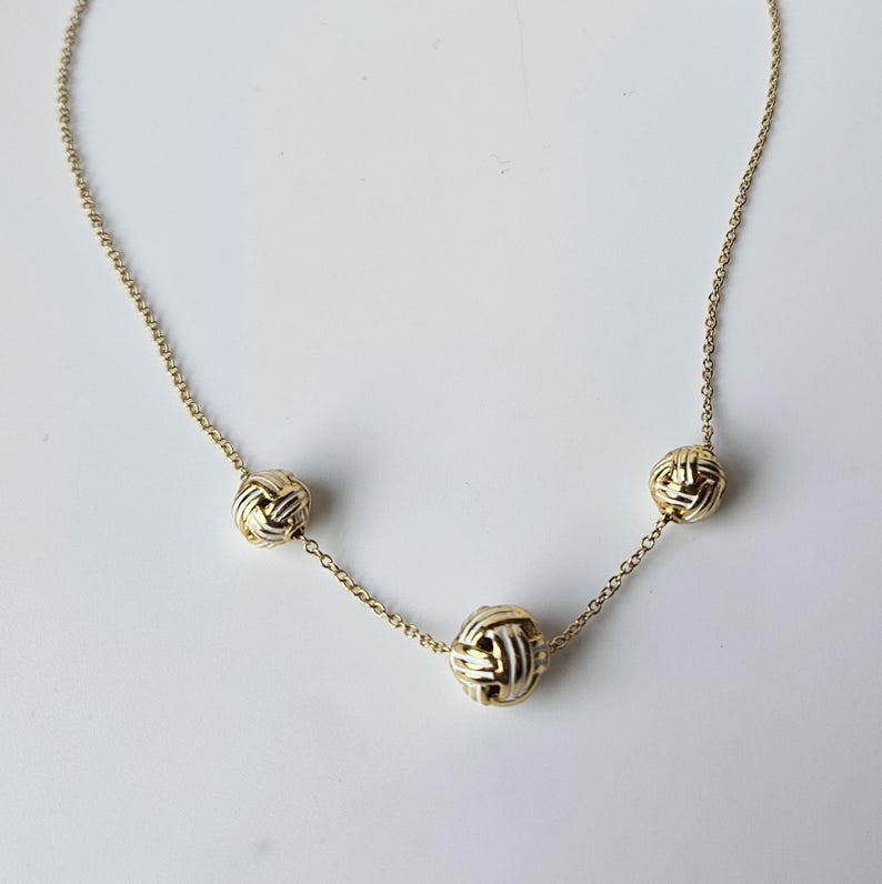 May include: A gold-toned necklace featuring three decorative knot-shaped pendants. The necklace has a delicate chain and the pendants have a woven design. The jewelry is displayed on a white background.