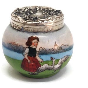 Könnte beinhalten: Eine kleine, runde Porzellan-Dose mit einem silbernen Deckel. Die Dose ist mit einer bemalten Szene eines Mädchens in einem roten Kleid und einer Gans auf einem grünen Feld verziert.