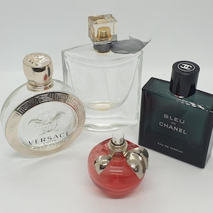 Può includere: Sono esposti quattro flaconi di profumo. Un flacone rotondo con dettagli dorati e la scritta "VERSACE EROS POUR FEMME" è accanto a un flacone quadrato trasparente. Sono visibili anche un flacone rotondo rosso e un flacone rettangolare blu scuro con "BLEU DE CHANEL".