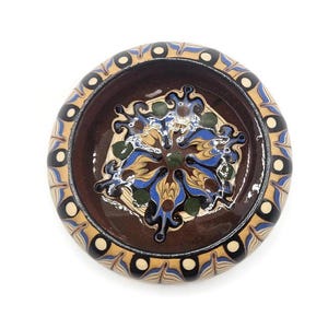 May include: A round, ceramic bowl with a brown interior and a decorative rim. The rim features a pattern of blue, tan, and black accents. The bowl's interior has a floral design in blue, yellow, and green.