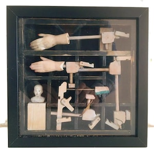 May include: A framed artwork with a black frame and glass front. Inside, wooden mechanisms and doll hands with red nail polish are arranged in a grid. A small bust is also visible, creating a surreal composition.
