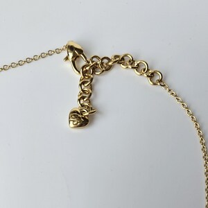 May include: Close-up of a gold-colored necklace clasp and chain. The necklace features a lobster claw clasp, a series of small, round chain links, and an extender chain with a heart-shaped charm. The jewelry is set against a white background.