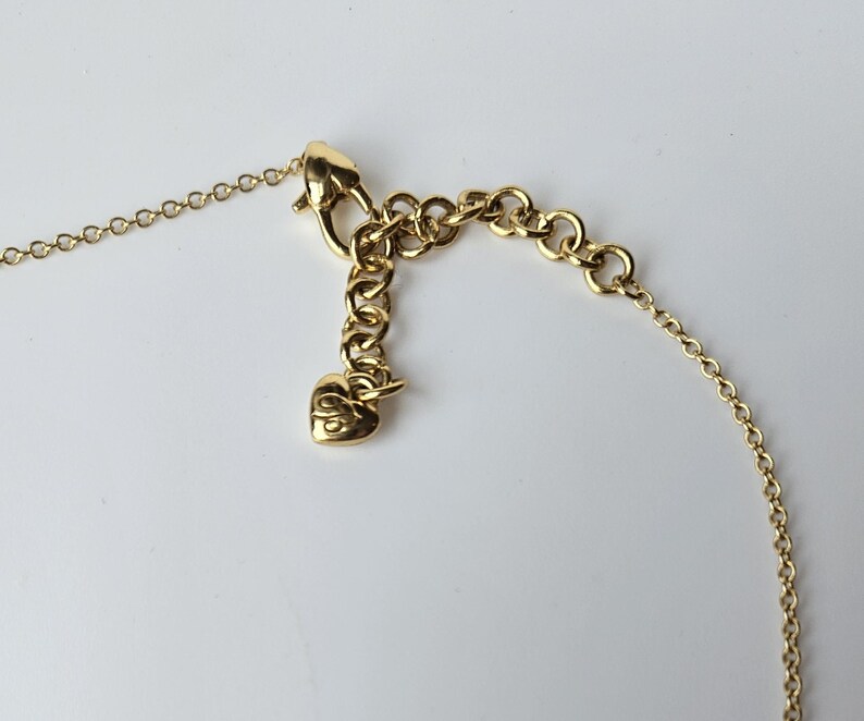 May include: Close-up of a gold-colored necklace clasp and chain. The necklace features a lobster claw clasp, a series of small, round chain links, and an extender chain with a heart-shaped charm. The jewelry is set against a white background.