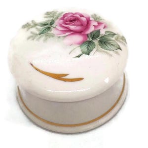 Könnte beinhalten: Kleine, runde Schmuckdose aus weißer Keramik mit Deckel. Der Deckel ist mit einer rosa Rose und grünen Blättern verziert. Ein goldfarbener Rand akzentuiert die Basis und den Deckel. Die Dose ist wahrscheinlich Vintage oder antik.