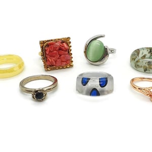 Op de afbeelding: Collectie ringen in diverse stijlen en kleuren. Inclusief een gele band, een gouden ring met rode stenen, een zilveren ring met een groene steen, een transparante ring met blauwe accenten en een roségouden ring met een paarse steen.