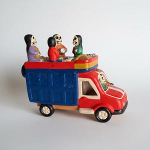 May include: A colorful, handcrafted ceramic truck featuring a red cab and blue cargo bed. The truck is adorned with skeleton figures, with one driving and others riding in the back. The truck has black wheels and is painted with bright colors.