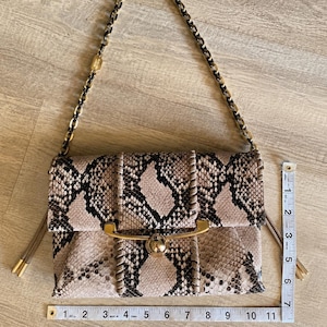 May include: A beige and black snakeskin-patterned handbag with a gold-tone chain strap and clasp. The bag has a rectangular shape with a flap closure and stitched detailing. The bag is approximately 11 inches wide.