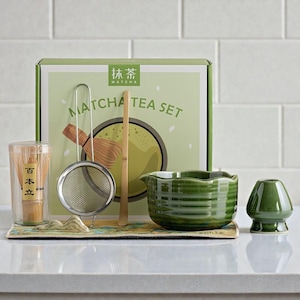 Puede incluir: Un juego de té Matcha que incluye un cuenco de cerámica verde, un batidor, un colador, una cuchara de bambú y un soporte para el batidor. El juego se exhibe sobre una alfombrilla decorativa frente a una caja con la inscripción "Matcha Tea Set".