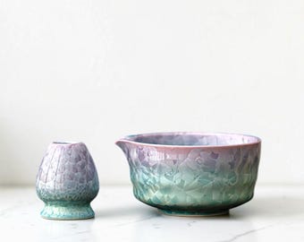May include: A ceramic matcha bowl and whisk set. The bowl is a gradient of green, blue, and purple with a textured crystalline pattern. The whisk is a similar color and texture. The set is designed for preparing matcha tea.
