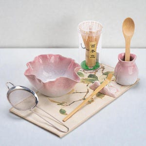 May include: A matcha tea set comprising a pink ceramic bowl, a bamboo whisk in a clear container, a wooden spoon in a pink ceramic holder, a bamboo scoop, and a stainless steel strainer. The set is displayed on a floral patterned mat.