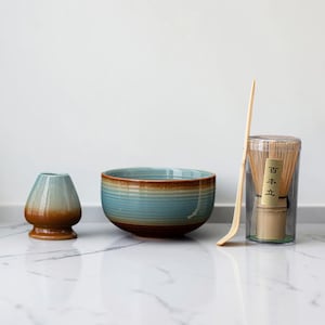 May include: A matcha tea set consisting of a ceramic whisk holder with a blue and brown gradient, a matching ceramic bowl with horizontal ridges, a long bamboo scoop, and a bamboo whisk inside a clear plastic case with the text 百本立.