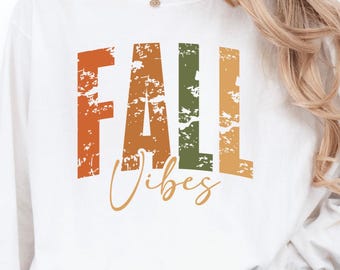 May include: White long-sleeved top featuring the text "FALL VIBES" in orange, green, and gold. The top is worn with blue jeans. A plant in a grey pot is in the background.