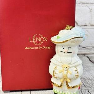 May include: A Lenox Santa Claus figurine with a white coat, holding a teddy bear. The figurine has a blue and gold hat and black boots. The red box behind the figurine has the Lenox logo and the words "American by Design."