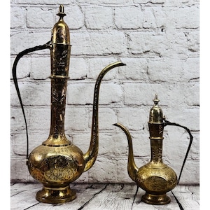 May include: Two ornate brass pitchers with intricate floral patterns. The larger pitcher has a tall, slender body with a long, curved spout and a handle. The smaller pitcher mirrors the design. Both are on a wooden surface.