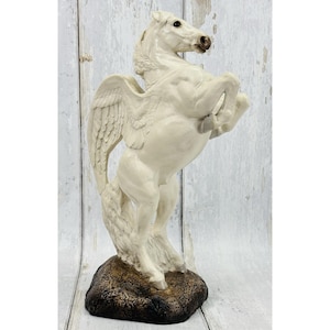 May include: A cream-colored Pegasus figurine depicted in a rearing pose with wings extended upward. The mythical horse stands on a textured, dark brown rock-like base. The sculpture features detailed musculature and a flowing mane and tail.