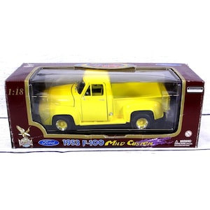 May include: A 1:18 scale model of a 1953 Ford F-100 Mild Custom pickup truck. The model is bright yellow and displayed in a clear plastic case with a maroon and brown cardboard box. The box features the Ford logo and the text "1953 F-100 Mild Custom".