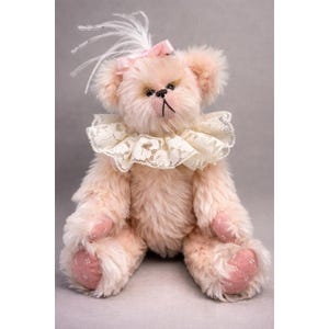May include: A plush, pink teddy bear with a frilly lace collar and a pink bow. The bear has a stitched nose and mouth, with pink paw pads. A small cluster of white feathers is attached to the top of the bear's head.