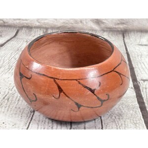 May include: A round, terracotta-colored ceramic bowl with a dark rim and black, curved line designs. The bowl has a wide opening and a smooth, unglazed surface. The design is reminiscent of Native American pottery.