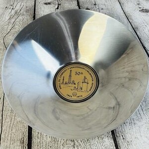 May include: A silver-toned metal ashtray with a circular design. The center features a gold-colored emblem with the text "Shell Oil Company Anniversary - Wilmington Refinery" and the years "1923-1973". The ashtray is on a wooden surface.