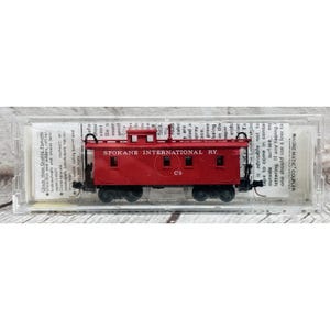 May include: A red model train caboose with the text "SPOKANE INTERNATIONAL RY." and "C5" printed on the side. The model is in a clear plastic case. The caboose has black wheels and a detailed roof.