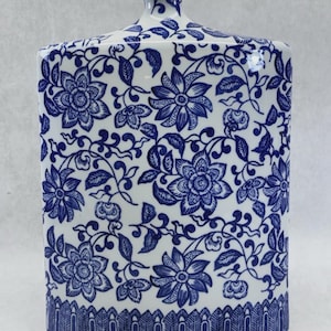 May include: A white ceramic jar with a blue floral pattern. The jar has a rectangular shape with a lid and a narrow neck. The floral design covers the entire surface of the jar, with a detailed border at the base.