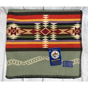 May include: A folded wool blanket featuring a geometric pattern in red, orange, purple, green, black, and cream. A blue Pendleton Beaver State tag is attached, along with a smaller blue label. The blanket has a black whip-stitched edge.