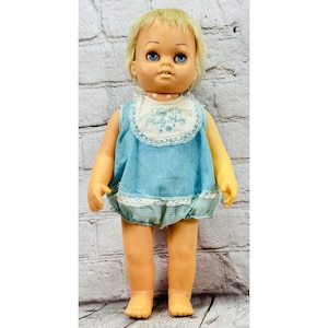 May include: A doll with blonde hair and blue eyes wearing a light blue sleeveless top with a white lace-trimmed bib featuring the embroidered text Tiny Chatty Baby in blue, paired with matching light blue and white striped bloomers with lace trim.
