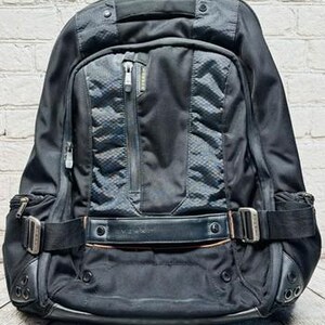 May include: Black backpack with multiple compartments, zippers, and adjustable straps. Features a textured front panel and a top handle. Includes silver-colored metal buckles and accents. The bag is designed for carrying various items.