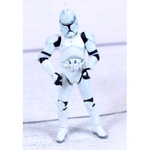 May include: A white plastic action figure of a clone trooper soldier wearing full body armor with black joint details and a helmet featuring a T-shaped visor. The figure stands in a neutral pose with articulated limbs and a small antenna on top.