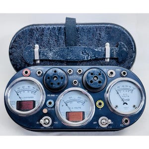 May include: A vintage, dark blue electronic testing device with multiple gauges and dials. The device features a black carrying case and several circular meters labeled "Milliamperes," "D.C. Volts," and "A.C. Volts." The device has a series of knobs and switches.