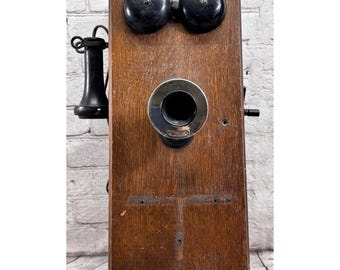 May include: Antique wooden wall phone with a dark finish. Features two black bells at the top, a circular mouthpiece, and a black handset on the left side. The phone has a vintage aesthetic.