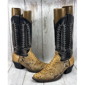 May include: A pair of cowboy boots featuring a textured, tan and brown snakeskin-patterned leather foot and a tall black leather shaft. The black shaft is decorated with intricate, light-colored stitching in a sharp, geometric flame-like pattern.