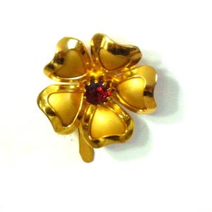 May include: A gold-toned flower-shaped brooch with five petals and a small, round, red gemstone at the center. The brooch has a glossy finish and is set against a white background.