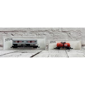 May include: Two model train cars in clear plastic cases. One is a gray flatcar with two containers labeled "SEA LAND". The other is an orange tanker car with "HOOKER" printed on it. Both are on a wooden surface.
