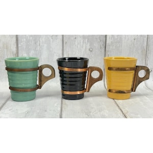 May include: Three ceramic mugs in light green, black, and yellow, each featuring horizontal ridges and a dark brown wooden handle attached by two thin, horizontal metal bands that wrap around the exterior of the mug body.