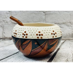 May include: A round ceramic bowl with a matching spoon. The bowl features a cream-colored top with a geometric pattern and brown dots. The lower half is decorated with brown and dark green leaf-like designs.