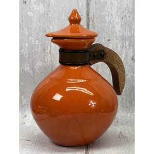 May include: A glossy orange ceramic pitcher with a matching lid featuring a pointed finial. The pitcher has a rounded body and a curved wooden handle attached to the neck with a dark metal band secured by two visible rivets on the side.
