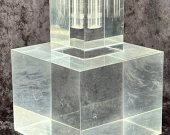 May include: A clear acrylic candle holder with a square base and a smaller square top. The top has two cylindrical holes for candles. The holder is made of thick, transparent material, and the edges are sharply defined.