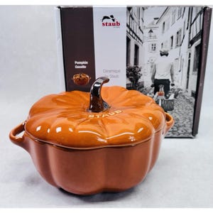 May include: An orange ceramic pumpkin cocotte with a silver handle and the word "STAUB" embossed on the lid. The cocotte is in front of a box with the words "Pumpkin Cocotte" and a black and white image of a street scene.