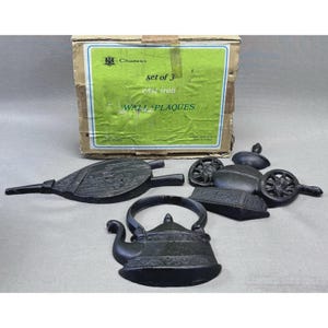 May include: A set of three black cast iron wall plaques featuring a bellows, a tea kettle, and a cart with wheels. The items are displayed in front of a cardboard box with a green label that reads Set of 3 cast iron WALL PLAQUES and Chadwicks.