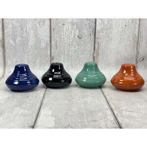 May include: Four ceramic salt and pepper shakers in a rounded, bell-like shape. The set includes one dark blue, one black, one light green, and one orange shaker, each featuring a smooth, glossy finish and a single hole at the top.
