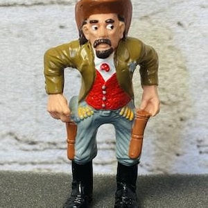 May include: A small figurine of a cowboy with a brown hat, green jacket, red vest, and blue pants. The cowboy has a mustache and is holding two brown gun holsters. Black boots complete the outfit.
