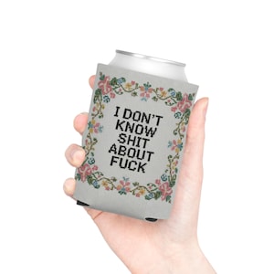 May include: A gray can cooler with the text "I DON'T KNOW SHIT ABOUT FUCK" in black. The cooler is decorated with a floral cross-stitch design in pink, blue, and green. The cooler is designed to keep beverages cold.