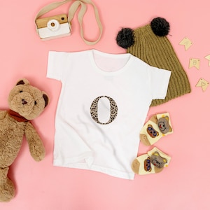 Pode incluir: Uma camiseta branca com a letra "O" estampada em leopardo &eacute; exibida em uma superf&iacute;cie rosa, junto com um ursinho de pel&uacute;cia, um gorro de malha com pompons e meias com desenhos de ursos. Uma c&acirc;mera de brinquedo e pequenas faixas de madeira tamb&eacute;m est&atilde;o presentes.