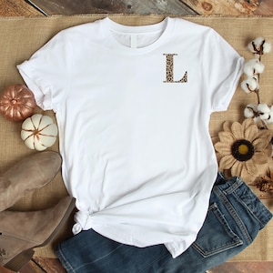 Pode incluir: Uma camiseta branca com a letra "L" com estampa de leopardo no peito. A camisa &eacute; estilizada com jeans azul, botas marrons e ab&oacute;boras e flores decorativas. A camisa &eacute; feita de um material macio.