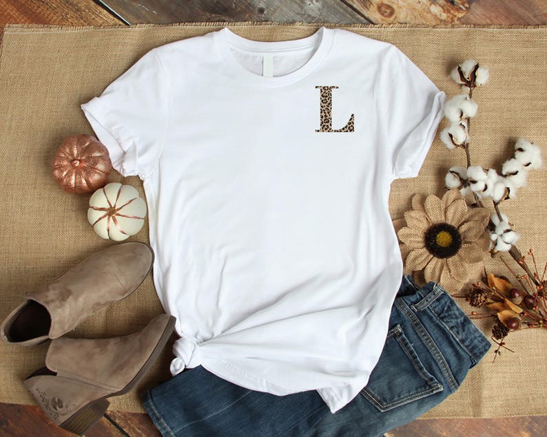 Pode incluir: Uma camiseta branca com a letra "L" com estampa de leopardo no peito. A camisa &eacute; estilizada com jeans azul, botas marrons e ab&oacute;boras e flores decorativas. A camisa &eacute; feita de um material macio.