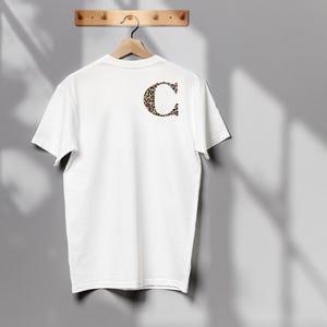 Pode incluir: Uma camiseta branca pendurada em um cabide de madeira. A parte de tr&aacute;s da camiseta apresenta uma grande letra "C" com estampa de leopardo. A camiseta &eacute; exibida contra uma parede cinza.