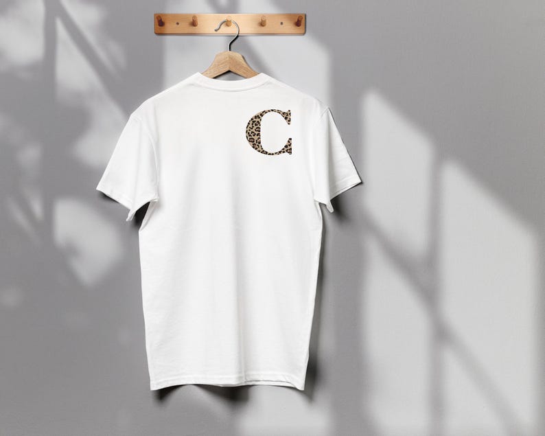 Pode incluir: Uma camiseta branca pendurada em um cabide de madeira. A parte de tr&aacute;s da camiseta apresenta uma grande letra "C" com estampa de leopardo. A camiseta &eacute; exibida contra uma parede cinza.