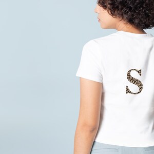Pode incluir: Camiseta branca com um "S" estampado com estampa de leopardo nas costas. A camiseta de manga curta tem gola redonda. A estampa de leopardo &eacute; uma mistura de manchas marrons e pretas. O fundo &eacute; azul claro.